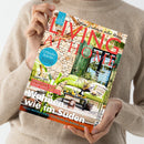 Person in beigem Strickpullover hält das Magazin „LIVING AT HOME“ vor neutralem Hintergrund; Titeltext in Rot mit Unterzeile „Mein Zuhause voller Ideen“. Auf dem Cover ein Außenbereich mit Steinwand, grüner Holztür, grünem Sofa und Couchtisch. Sichtbare Texte: „Urlaubsküche – Salat, Steak, Eis“, „Wohnen wie im Süden“, „Sommerliche Ideen für den Tisch“, „Citytrip München“ und „Tolle Inspirationen von Palma bis Buenos Aires“.