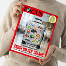 Eine Person in beigefarbenem Strickpullover hält eine Ausgabe des Magazins „FOCUS“. Das Titelbild zeigt einen silberfarbenen Rollkoffer mit Aufklebern, darunter ein Flugzeug, Autos, eine Weltkugel, ein Vulkan mit „BOOM!“, Palmeninsel, Gebirge sowie eine Person mit roter Krawatte, die nach rechts zeigt. Haupttitel: „ANGST UM DEN URLAUB“, Untertitel: „Die 30 wichtigsten Fragen zum Krisen-Sommer 2026“.
