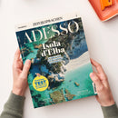 Titelblatt des Magazins „Adesso“ der Reihe „Zeit Sprachen“, Ausgabe 7/25, mit der Überschrift „Isola d’Elba – So lebt es sich auf der coolsten Insel Italiens!“. Das Cover zeigt eine Luftaufnahme der Küste von Elba mit türkisblauem Wasser, hellen Felsen und einem Strand. Unten links gelber Kreis mit der Aufschrift „Einstufungs-Test: Wie steht es um Ihr Italienisch?“. Gehalten von zwei Händen über hellem Hintergrund.
