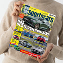 Person in beigem Strickpullover hält ein Magazin mit gelbem Titelbalken „Auto Bild sportscars“. Das Cover zeigt mehrere Sportwagen-Fotos, im Mittelpunkt ein grüner Audi RS 3 mit der Überschrift „Das Beste zum Schluss – Audi RS 3 competition limited im Test“. Weitere sichtbare Texte: Themen zu Technik, Gebrauchtwagen, Corvette ZR1, BMW M Concept, Porsche-Spezialist und Mini Cooper C.