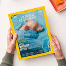 Titelblatt der Zeitschrift „National Geographic“, Ausgabe Juni 2025, mit gelbem Rand und zentralem Foto eines neugeborenen rosa Ferkels, das in blauen Latexhandschuhen auf hellblauer Unterlage gehalten wird; Hauptschriftzug: „DIESES SCHWEIN KANN MENSCHEN RETTEN“, zusätzliche Texte zu Themen wie Organtransplantation, Konflikte um Biber, Pluto-Debatte, Inseln in der Wüste und LGBTQ+. Hände in grünem Strickärmel halten das Magazin.