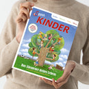 Zeitschrift mit dem Titel „Servus Kinder“ in roter Schrift und Unterzeile „Spielen | Staunen | Rätseln | Wissen“, gehalten von einer Person in beigefarbenem Strickpullover. Covermotiv: große Illustration eines Baumes mit spielenden Kindern, darunter grüne Wiese mit Kuh und Zaun, unten Text „Das Abenteuer deines Lebens – Tausend Ideen für große und kleine Entdecker“.