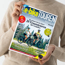 Person in beigefarbenem Pullover hält das Magazin „e‑bikeTOUREN MAGAZIN“ (Ausgabe 02‑26 Frühling/Sommer) vor neutralem Hintergrund. Auf dem Cover fahren zwei Personen mit Helmen auf schwarzen E‑Bikes durch eine grüne Wiesenlandschaft, darüber Texte wie „20 Traumtouren für Genießer“, „Klassiker Donau‑Radweg“, „E‑Bike‑Motoren“ und „Märchenhafte Mittelgebirge“. Farbtöne in Blau, Gelb, Weiß und Rot dominieren das Layout.