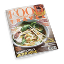 Titelbild des Magazins „Food and Travel“ in orangefarbener Schrift mit dem Untertitel „Deutsch“. Im Vordergrund eine große Schüssel Nudelsuppe mit Gemüse, Kräutern, Knoblauchscheiben und zwei Holzstäbchen, von schräg oben fotografiert. Text auf dem Cover: „Rezepte für den Frühling“, „Irisch backen leicht gemacht“, „Neue Safari-Lodges“, „Stadthotels unter 200 €“, „Malaysias Gourmetinsel Langkawi“, „Kulinarik-Hotspot Cadiz“, „Cordoba“ und „Skifahren im Spätwinter“.