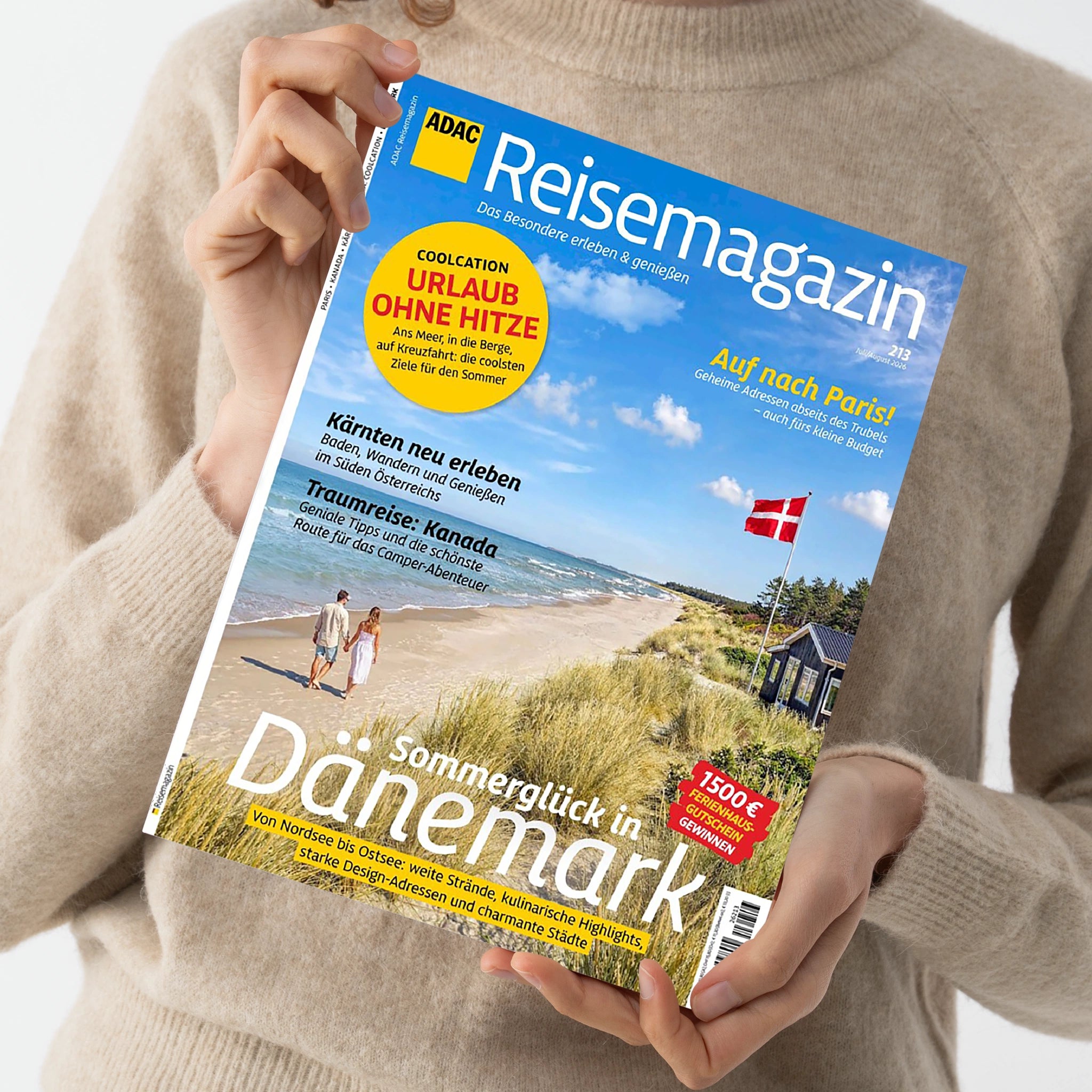 Person in beigem Strickpullover hält das „ADAC Reisemagazin“ mit blauem Titelbanner und gelbem Logo. Covermotiv zeigt einen Sandstrand in Dänemark mit Dünen, Meer, blauem Himmel, roter dänischer Flagge und einem Haus am rechten Bildrand. Hauptüberschrift: „Sommerglück in Dänemark“. Weitere Texte: „URLAUB OHNE HITZE“, „Kärnten neu erleben“, „Traumreise: Kanada“, „Auf nach Paris!“.