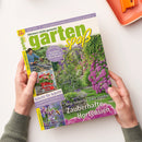 Titelbild der Gartenzeitschrift „gartenspaß“ Juni 2025 mit dem Untertitel „Gärtnern leicht gemacht“. Zentrale Abbildung eines blühenden Gartens mit lila Hortensien und Rosenbogen, umgeben von weiteren Pflanzen. Textaussagen wie „Zauberhafte Hortensien“, „Praktisch & naturnah“, „Schritt für Schritt“ und „Bienenmagnete“ sind sichtbar. Eine Person hält das Magazin mit beiden Händen über einer hellen Tischfläche, daneben eine orange Schale.