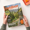 Magazintitel „outdoor“ in großer oranger Schrift auf einem Cover mit Berg- und Waldlandschaft; zwei wandernde Personen auf einem Pfad, umgeben von grünen Bäumen und Felsen. Textaufschriften: „TOP-EQUIPMENT FÜRS CAMPING“, „Abenteuer Jotunheimen“, „Bayerns stille Winkel“, „IM CHECK: SOFTSHELLS“, „Schwarzwald entdecken“ und weitere Themen zu Wandern und Ausrüstung. Zwei Hände halten das Magazin über einem hellen Tisch mit orangefarbener Schale.