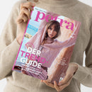 Person in beigem Strickpullover hält ein Exemplar der Zeitschrift „petra“ mit pinkfarbenem Schriftzug. Auf dem Titel ist eine weibliche Person in rosa Zopfpullover mit heller Umhängetasche vor sandfarbenem Felsen zu sehen. Texte auf dem Cover: „DER TREND-GUIDE“, „TRAUMKLEIDER“, „IT-BAGS“, „SUMMER-SHOES“, „BEAUTY-HIGHLIGHTS“, „BEST OF GERMANY“ und „STIL & HALTUNG“.