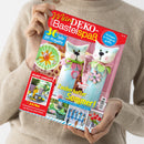 Frau in beigem Strickpullover hält eine Bastelzeitschrift mit dem Titel „Mein DEKO und Bastelspaß“. Auf dem bunt gestalteten Cover stehen Schlagzeilen wie „30 tolle DIY-Projekte“, „Zauberhafter Sommer!“, „Selbst gemacht!“ und „Extra: Farbenfrisch & sommerlich mit Zitronen dekorieren“. Abgebildet sind zwei Filzkatzen in Pastellfarben, Papierblumen, Bastelideen und sommerliche Deko-Projekte vor rosafarbenem Hintergrund.