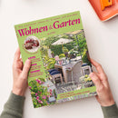 Magazin „Wohnen & Garten“ Juni 2025 mit grünem Titelblatt und pinkfarbener Schrift, gehalten von zwei Händen über hellem Hintergrund; zentrales Titelbild zeigt gedeckten Gartentisch mit Rattanmöbeln, Blumen und Sonnenschirm. Sichtbare Texte: „Die schönsten Ideen für drinnen und draußen“, „Willkommen im Sommersalon“, Themenrubriken „Wohnen“, „Garten“, „Dekorieren“ sowie Hinweis auf „Köstliche Rezepte mit Kirschen“.