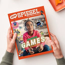 Titelblatt des Magazins „Dein SPIEGEL“ mit dem Untertitel „Durch Wissen wachsen“. Im Mittelpunkt ein Kind mit Kopfhörern und Spiele-Controller, umgeben von bunten Spielfiguren und digitalen Symbolen. Große grüne und gelbe Schrift: „Die besten Games – für euch getestet“. Oben links kleine Bilder mit Überschriften „Die Weltenlenkerin“ und „Was startet, wenn die Rakete auf...“. Umschlag mit orangefarbenem Rand, neutraler Hintergrund.