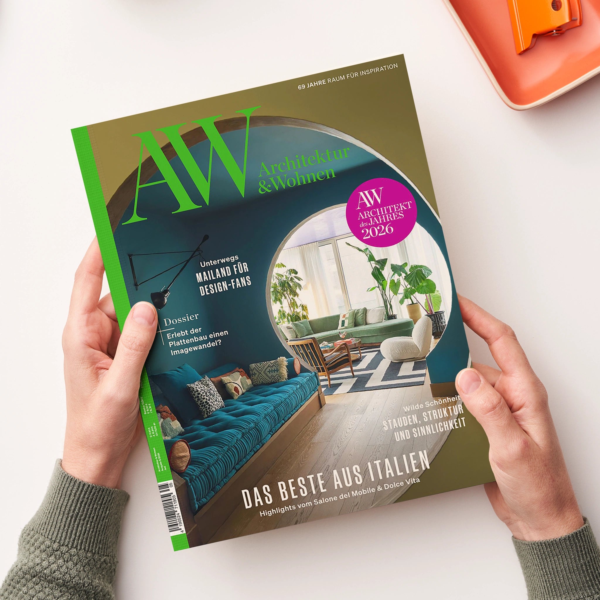 Titelblatt der Zeitschrift „AW Architektur & Wohnen“ mit großem grünem „AW“-Logo auf olivgrünem Hintergrund, Unterzeilen in Türkis zu Themen wie „Mailand für Design-Fans“ und „Das Beste aus Italien“. Mittig ein rundes Fenstermotiv mit Blick auf modern eingerichtete Wohnräume in Grün- und Holztönen, oben rechts pinkfarbener Kreis mit Text „AW Architekt des Jahres 2026“, gehalten von zwei Händen über hellem Tisch.