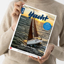 Helle, langärmlig gekleidete Person hält das Magazin „Yacht“ in beiden Händen vor neutralem Hintergrund; das Cover zeigt ein Segelboot mit braunen Segeln auf bewegter Meeresoberfläche bei Sonnenuntergang. Sichtbare Texte: Magazintitel „Yacht“, Überschriften „Ostsee“, „Endlich Eigner“, „Kleinkreuzer-Refit“, „Ab auf die Welle!“ sowie blauer Kreis mit „Segel-Influencer – Von Bootsbau bis Abenteurer: Wie sich Segler auf Social Media zeigen“.