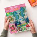 Titelblatt der Bastelzeitschrift „Mein DEKO- und Bastelspaß“ mit zwei blauen, glitzernden Seepferdchen als Hauptmotiv auf hellblau-grünem Hintergrund, gelber Schrift „Hallo Sommer!“ und Texten wie „30 tolle DIY-Projekte“ und „SELST GEMACHT!“. Im unteren Bereich Bilder von Bastelideen, darunter Blüten, Kerzen und Gartenfiguren. Zwei Hände halten das Heft vor hellem Untergrund.