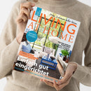 Helle Person in beigefarbenem Strickpullover hält eine Ausgabe des Magazins „Living at Home“. Titel in orange und türkis, mit Unterzeilen wie „Einfach gut eingerichtet“, „Spargelrezepte“, „Per Rad durchs Ruhrgebiet“, „Ein bunter Naturgarten“. Auf dem Cover ein modern eingerichtetes Wohnzimmer mit blauem Tisch, Sofas, Kissen und großen Fenstern mit Blick auf grüne Pflanzen.