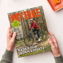 Titelblatt der Zeitschrift „MY BIKE“ mit großem orangefarbenem Schriftzug oben. Abgebildet ist eine maskuline Person in roter Jacke, dunkler Hose und grauem Fahrradhelm, die ein graues E-Bike durch einen Wald fährt. Textausschnitte: „Räder für jeden Einsatz“, „20 Seiten Basiswissen zum Fahrradkauf“, „Frühjahrs-Check“, „Citytrip Lissabon“. Das Heft wird von zwei Händen auf heller Tischoberfläche gehalten.