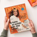 Magazin „DONNA“ mit orangefarbenem Titelhintergrund, darauf eine lächelnde weibliche Person mit lockigem dunklem Haar und weißer Jacke. Hauptüberschrift: „Es kann so schön sein, das Leben“. Weitere sichtbare Texte: „Amore! Italienische Rezepte, ganz easy“, „Gen D – Generation DONNA“, „Ich will nicht jünger aussehen“, sowie Themen zu Liebe, Fitness, Promis und Wandern. Hände halten das Heft über hellem Tisch mit orangefarbenem Objekt rechts oben.
