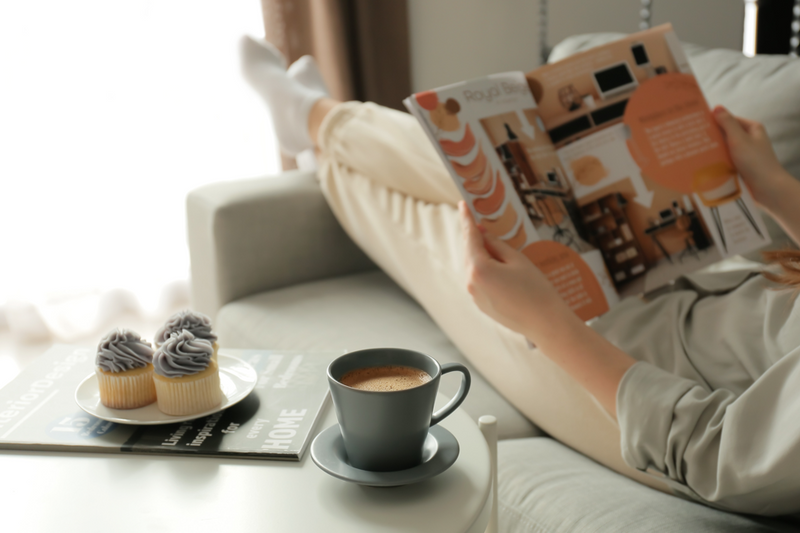 Person liest auf dem Sofa ein Magazin über Innendesign, daneben eine Tasse Kaffee und Cupcakes.