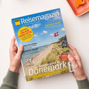 Magazintitel „ADAC Reisemagazin“ mit blauem Hintergrund und Foto einer Küstenlandschaft mit Dünen, Sandstrand, Meer und Haus mit dänischer Flagge; im Vordergrund zwei gehende Personen. Gelbe, blaue und weiße Überschriften unter anderem: „Sommerglück in Dänemark“, „URLAUB OHNE HITZE“, „Traumreise: Kanada“ und „Auf nach Paris!“. Gehalten von zwei Händen vor hellem Hintergrund, rechte obere Ecke der Seite leicht angehoben.