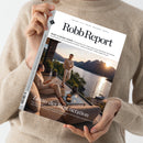 Person in beigefarbenem Strickpullover hält ein Magazin mit dem Titel „Robb Report“ und dem Untertitel „Body & Mind Issue“. Auf dem Cover sitzt eine Person auf einem Sofa, eine weitere steht barfuß davor; beide tragen helle Kleidung. Im Hintergrund ist eine Seeterrasse mit Topfpflanzen, Glasgeländer und Berglandschaft bei Sonnenuntergang zu sehen. Textausschnitt: „Luxus der Regeneration“.