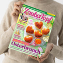 Eine Person in beigefarbenem Strickpullover hält ein Magazin mit dem Titel „ZauberTopf – Rezepte für Thermomix“ in pink-lila Schrift. Das Cover zeigt fünf goldbraune, herzförmige Hefegebäcke auf weißem Untergrund, daneben ein Glas mit oranger Konfitüre und ein rosa Tulpenblatt. Sichtbare Textelemente: „Liebevolle Ideen: Osterbrunch“, „Hollandaise“, „Eierlikör“, „Alles strahlt!“, „NEU: Einfache Routinen für mehr Energie“.