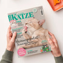 Titelbild der Zeitschrift „Geliebte Katze“, Ausgabe Mai 2025, mit einer beigen Katze, die sich die Pfote leckt, auf grauem Hintergrund. Textelemente: „Herausgeputzt! Die beste Pflege-Routine für jeden Fell-Typ“, „Kitten-Quiz – Testen Sie Ihr Wissen rund um Katzenbabys“, „Tierschutz – Das geheime Leben der Straßenkatzen“, „5 frische Frühlings-Ideen“. Zwei Hände in grünem Pullover halten die Zeitschrift über hellem Untergrund.