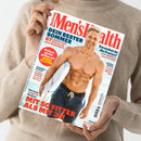 Person in beigefarbenem Strickpullover hält ein Exemplar des Magazins „Men’s Health“. Das Cover zeigt einen muskulösen, lächelnden Mann mit Surfbrett am Strand, bekleidet mit dunkelblauer Badehose. Haupttitel in roter Schrift: „Men’s Health“, weitere Schlagzeilen u. a. „Dein bester Sommer“, „Mit 50 fitter als mit 35!“, „Einfach genial grillen mit Jamie Oliver“ und Themen zu Fitness, Ernährung und Dating.