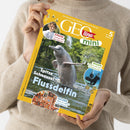Person in beigefarbenem Pullover hält eine Ausgabe des Kinderwissensmagazins „GEOlino mini“ mit gelbem Rand und Titelthema „Spitze Schnauzel! Flussdelfin“. Auf dem Titelbild springt ein grauer Flussdelfin aus dem Wasser vor grünem Hintergrund. Sichtbare Textfelder: „KINDER Carissa bei den Bienen“, „WISSEN Komm mit auf Schatzsuche“, „SUCHEN Alles in toller Unordnung“, Angaben „ab 5 Jahren“ und „Nr. 6/2023“.