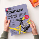 Titelblatt der Zeitschrift „Finanzen“ von Stiftung Warentest, gehalten von zwei Händen über heller Oberfläche. Lila Hintergrund, im unteren Bereich ein gelbes Fahrrad mit schwarzem Sattel und angeschlossenem Fahrradschloss. Schwarze Überschriften u. a. „Top-Schutz für Ihr Rad“, „Versichern Sie E-Bike und Fahrrad gegen Diebstahl“ sowie roter Kreis mit weißem Text „Plus: Die besten Fahrradschlösser“.