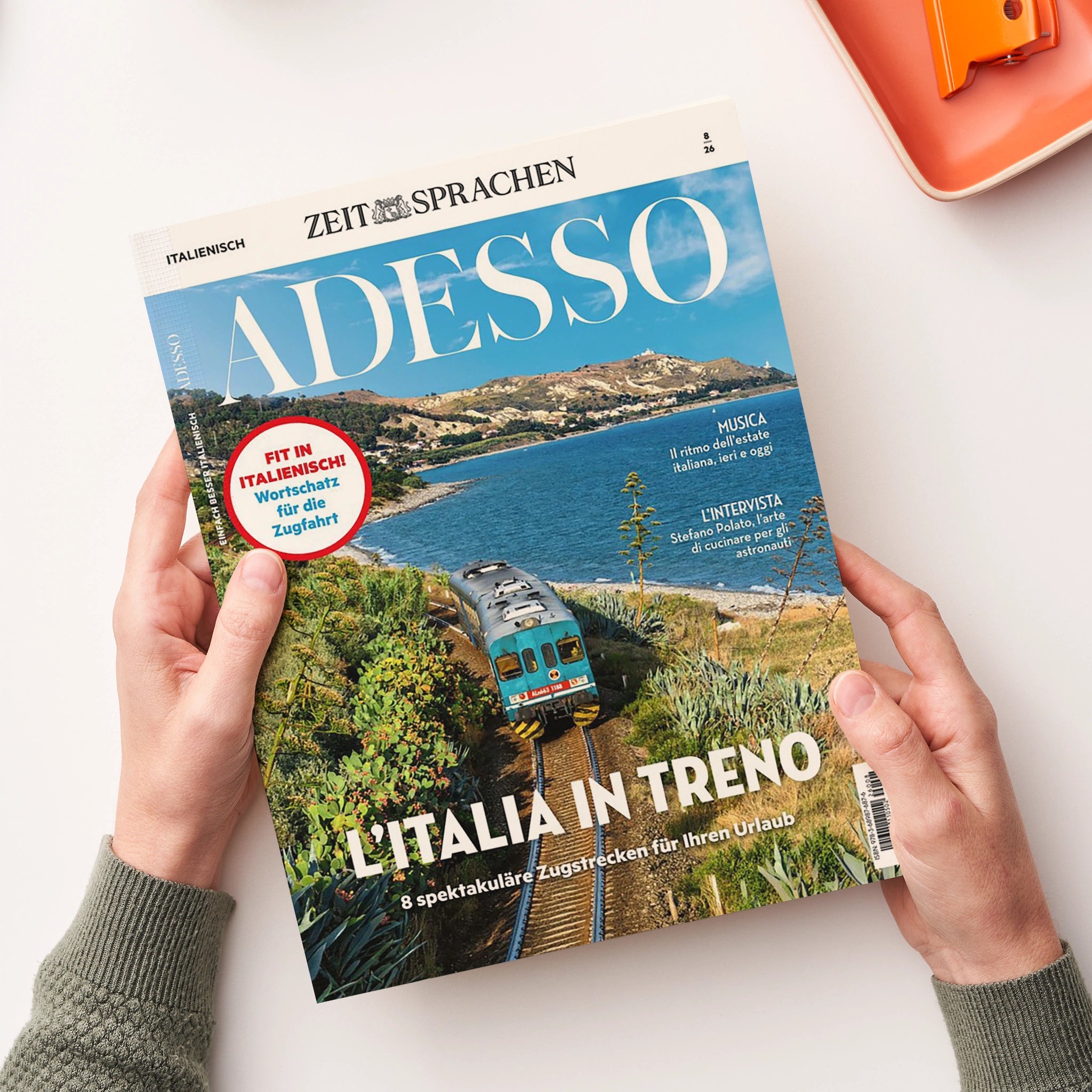 Magazin „ADESSO“ aus der Reihe „ZEIT SPRACHEN“, gehalten von zwei Händen über hellem Untergrund; Titelbild zeigt eine grün-blaue Regionalbahn auf einer Küstenstrecke mit Meer, Hügeln und Vegetation im Hintergrund. Aufdrucke u. a.: „FIT IN ITALIENISCH! Wortschatz für die Zugfahrt“, „L’ITALIA IN TRENO“, „8 spektakuläre Zugstrecken für Ihren Urlaub“, „MUSICA“ und „L’INTERVISTA“.