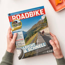 Titelblatt der Zeitschrift „ROADBIKE“ mit rotem Schriftzug auf blauem Hintergrund; im Hauptbild fahren drei Personen auf Rennrädern eine asphaltierte Bergstraße entlang, rechts eine Steinmauer, links Berglandschaft mit grün-braunen Hängen und blauem Himmel. Gelbe und weiße Textfelder mit Aufschriften wie „LEICHT & SCHNELL“, „GUT & GÜNSTIG“ und weiteren Themen zu Tests, Equipment und Italien-Touren. Hände halten das Magazin über hellem Untergrund.
