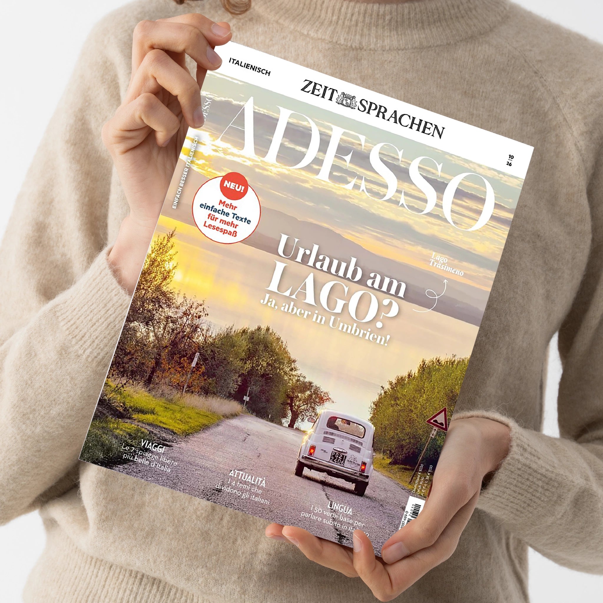 Hochformatiges Magazin „ADESSO“ von „ZEIT SPRACHEN“ in den Händen einer Person mit beigem Strickpullover. Das Cover zeigt eine italienische Landschaft mit Straße, weißem Auto und Verkehrsschild sowie die Texte „ITALIENISCH“, „EINFACH BESSER ITALIENISCH“, „Urlaub am LAGO? Ja, aber in Umbrien!“, „NEU! Mehr einfache Texte für mehr Lesespaß“, „VIAGGI“, „ATTUALITÀ“ und „LINGUA“.