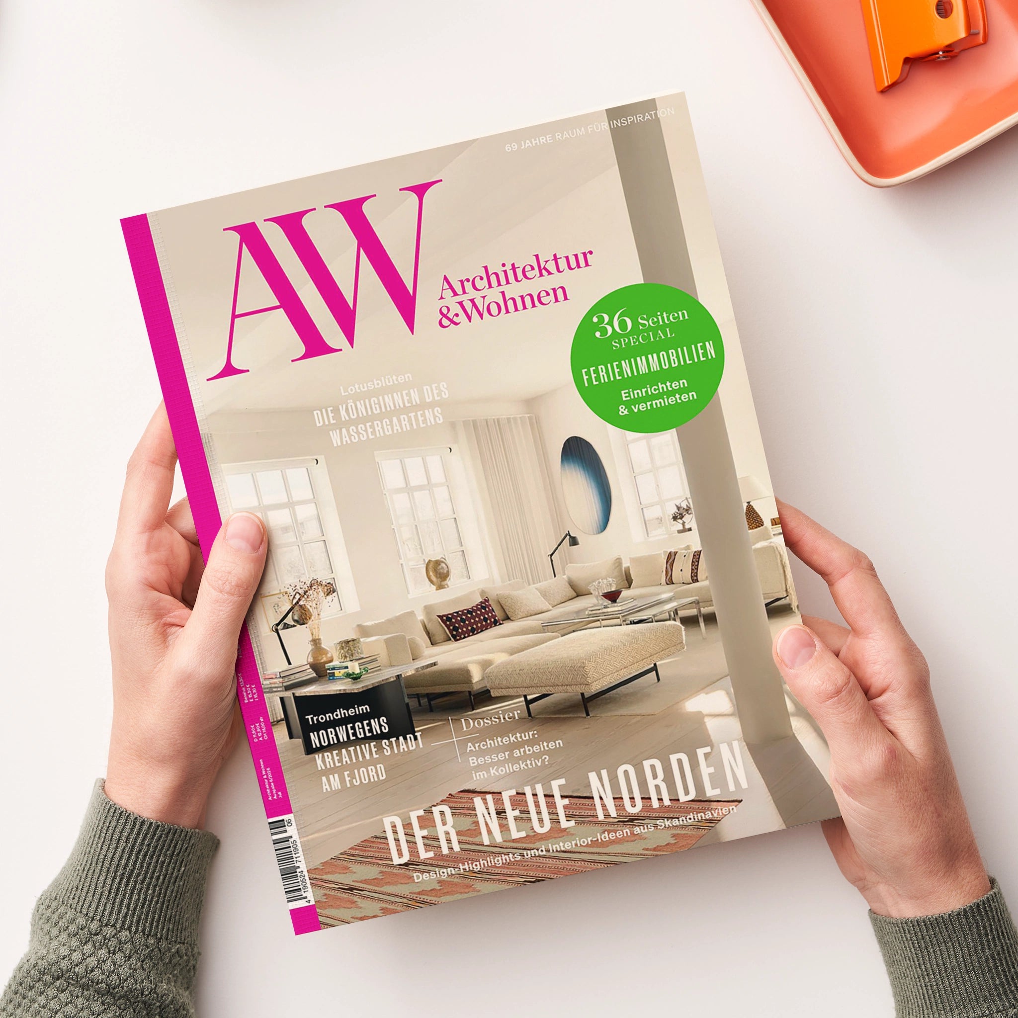 Von zwei Händen gehaltenes, schräg liegendes Magazin „AW Architektur & Wohnen“ mit cremefarbenem Cover, pinkem Schriftzug und Foto eines hellen Wohnzimmers. Sichtbare Texte: „69 Jahre Raum für Inspiration“, „36 Seiten Spezial Ferienimmobilien Einrichten & vermieten“, „Der neue Norden“ und „Design-Highlights und Interior-Ideen aus Skandinavien“; oben rechts liegt teilweise ein orangefarbenes Objekt.