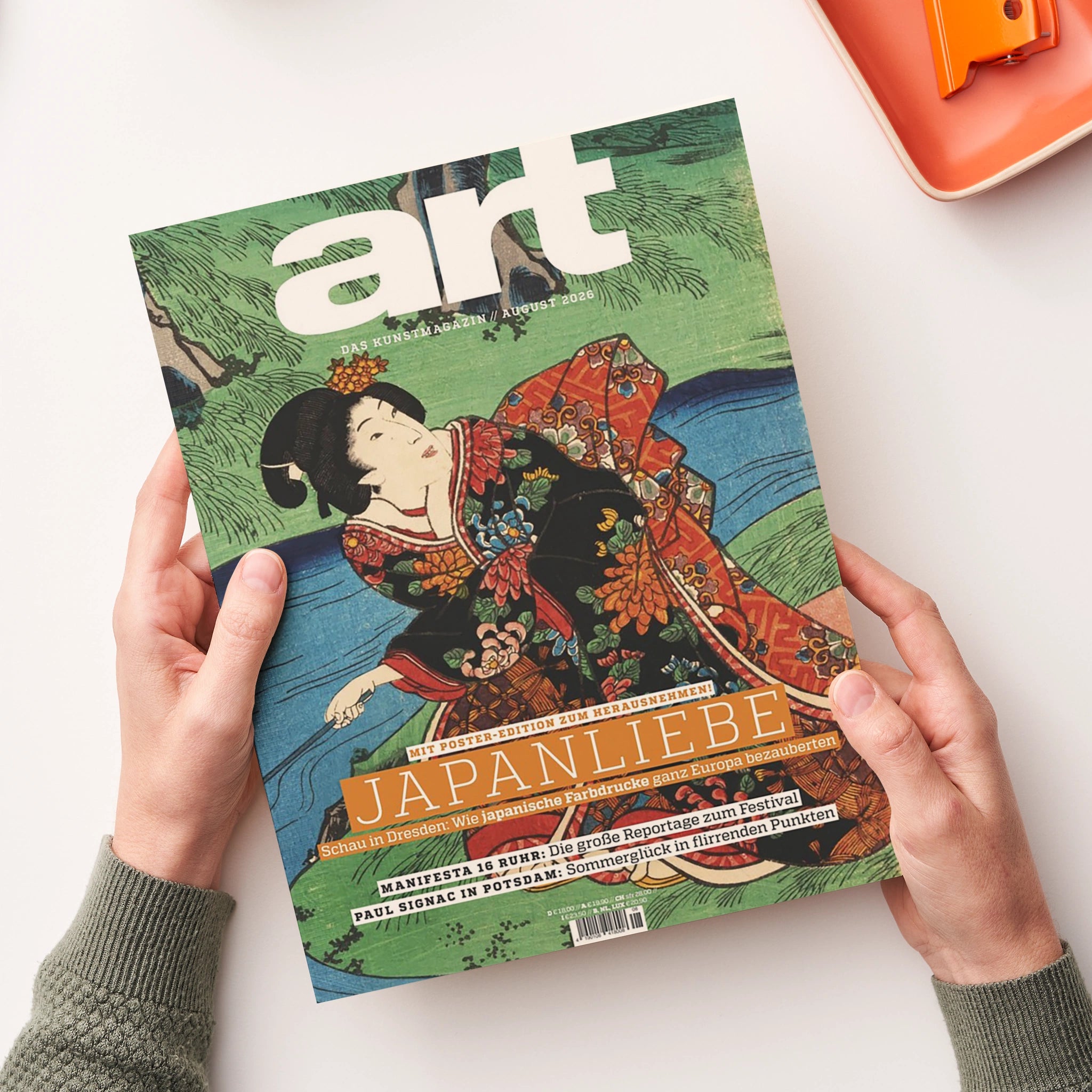 Aufgeschlagenes Kunstmagazin „art“ in schräger Draufsicht, von zwei Händen über weißer Fläche gehalten; Cover mit farbiger japanischer Holzschnittdarstellung einer Frau im dekorierten Kimono vor grüner Landschaft und blauem Wasser. Sichtbare Texte: „DAS KUNSTMAGAZIN // AUGUST 2026“, „JAPANLIEBE“, „MIT POSTER-EDITION ZUM HERAUSNEHMEN!“, „MANIFESTA 16 RUHR“ und „PAUL SIGNAC IN POTSDAM“.