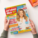 Hochformatige Zeitschrift „auf einen Blick“ liegt diagonal auf einer weißen Fläche und wird von zwei Händen gehalten. Das farbige Cover zeigt eine lächelnde blonde Frau mit weißer Bluse sowie die Texte „2 Hefte in einem! Ihre Zeitschrift für TV und Ratgeber“, „Alle TV-Höhepunkte“, „Finanz-Tipps für jedes Alter“ und „Sommerlicher Rouladen-Genuss“; oben rechts liegt ein orangefarbenes rechteckiges Objekt.