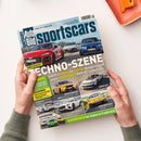 Schräg gehaltenes Magazin „sportscars BILD Auto“ auf weißer Fläche, von zwei Händen mit graugrünen Ärmeln. Das Cover zeigt rote, weiße und blaue Sportwagen sowie weitere Fahrzeugabbildungen und die Texte „TECHNO-SZENE“, „VERGLEICHSTEST“, „Audi RS 5 HYBRID und ELEKTRO Hyundai Ioniq 6 N gegen VERBRENNER BMW M3 Competition“ und „HAUSBESUCH bei Alpine in Frankreich“.