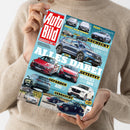 Eine Person im beigefarbenen Strickpullover hält schräg eine Zeitschrift mit blau-türkisem Cover und rotem „AUTO BILD“-Logo. Abgebildet sind zahlreiche Fahrzeuge; lesbare Texte sind „EUROPAS NR. 1“, „BENZIN ODER STROM? PEUGEOT 308 SW“, „TEST Hyundai Ioniq 6 N“, „GECHECKT GEBRAUCHTE KOMPAKT-SUV“, „GETESTET MERCEDES CLA GEGEN BMW 220“, „GEFAHREN AUDI Q7“, „GEBRAUCHT Mazda CX-60“, „DACHZELTE IM TEST“ und „FASZINATION PORSCHE 964“.