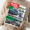 Person im beigen Strickpullover hält ein schräg fotografiertes Magazin „auto motor sport“ mit rotem Titelbereich und zahlreichen Fahrzeugabbildungen. Lesbare Überschriften sind „Was die Hersteller planen. Und was kommt NEUHEITEN 2027“, „Fährt Audi wieder vorne?“, „Vergleichstest BILLIGER TESLA“, „Travel-Guide ALPENÜBERQUERUNG“ und „Gebrauchtwagen MERCEDES S-KLASSE“; daneben stehen kleinere Fahrzeugbezeichnungen und ein Barcode.