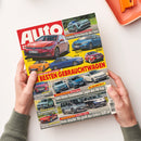 Aufgeschlagene Automobilzeitschrift mit großem „auto“-Logo, zahlreichen Fotos roter, blauer, grüner und weißer Fahrzeuge sowie gelben und roten Überschriften, darunter „Die besten Gebrauchtwagen“, „Stärkster Serien-Porsche aller Zeiten“ und „Vergleichstest Familien-Vans“. Zwei Hände ohne erkennbare Geschlechtsmerkmale halten das Magazin über einer weißen Fläche; oben rechts liegt ein orangefarbenes Notizbuch mit Klammer.
