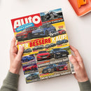 Titelblatt der Autozeitschrift „Auto“ in roten Großbuchstaben auf rotem Hintergrund, gehalten von einer Person mit grünem Strickpullover über hellem Tisch. Abgebildet sind mehrere Autos verschiedener Marken wie VW, Toyota, Hyundai, Alfa und Mercedes in unterschiedlichen Farben, dazu Schlagzeilen wie „DER BESSERE KAUF!“ und „Spannendes Mittelklasse-Duell“, mit weiteren Texten zu Vergleichen, Tests und Modellen.