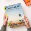 Zeitschrift „Robb Report – The New Luxury“, deutsche Ausgabe Nr. 38, wird von zwei Händen in graugrünen Ärmeln auf heller Oberfläche gehalten. Titelbild zeigt ein weißes Auto im Wüstenstaub mit Textfeld „WER BREMST, VERLIERT“ sowie kleineren Überschriften zu Themen wie Reisen, Design und Lifestyle. Linker Rand in hellgrün mit schwarzem Logo „R“. Orangefarbenes Tablett oben rechts im Anschnitt sichtbar.