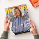 Titelblatt der Zeitschrift „freundin“ mit einer lächelnden weiblichen Person in hellblauer Bluse mit dunkelblauer Zierstickerei vor hellem Hintergrund. Aufschriften u. a.: „Leichter durchs Leben“, „Neue Frisur, neuer Look“, „Hormone besser verstehen“, „Wir lieben Romantik-Mode“ und „Die schönsten Ecken in Griechenland“. Gehalten von zwei Händen über weißer Oberfläche neben orangefarbenem Tablett.