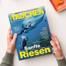 Titelblatt der Zeitschrift „TAUCHEN“ mit blauem Unterwasserfoto, das eine Person in Tauchausrüstung neben einem großen Mantarochen zeigt; gelbe Überschriften wie „Sanfte Riesen – Alles Wissenswerte über Mantas“, Themenrubriken zu „Reisewelten“, „Medizin“, „Tests & Tipps“ und „Kwazulu-Natal“; gehalten von zwei Händen über heller Tischoberfläche mit orangefarbenem Tablett im Hintergrund.