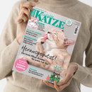 Person in beigem Strickpullover hält Zeitschrift mit dem Titel „Geliebte Katze“ in helltürkiser Schrift. Auf dem Cover leckt sich eine beige Katze selbst das Fell, daneben Texte wie „Herausgeputzt! Die beste Pflege-Routine für jeden Fell-Typ“, „Das große Krabbeln – Wirksamer Schutz vor Floh & Zecke“, „Plötzlich anders“, „Tierschutz – Das geheime Leben der Straßenkatzen“ und „So werden Sie zum Katzen-Liebling“.