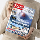 Person in beigefarbenem Pullover hält das Magazin „FLUG REVUE – Das Luft- und Raumfahrt-Magazin“, Ausgabe Juni 2025. Cover zeigt ein futuristisches Flugzeug unter der Überschrift „Der nächste US-Superfighter“, mit weiteren Texten „Boeing F-47: Erste Bilder“, „Was plant Airbus für sein nächstes Flugzeug?“ und kleineren Bildern von Flugzeugen, Cockpit und Messehalle, vor hellblauem Hintergrund.