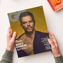Titelblatt der Zeitschrift „GQ Germany“ mit einem männlichen Modell vor gelbem Hintergrund, trägt dunkelviolettes Hemd mit geöffnetem Kragen. Text auf dem Cover: „DIE ODYSSEE MIT MATT DAMON“, „TOM HOLLAND & ROBERT PATTINSON“, „JEREMY POPE: DER STAR IN UNSERER PRIDE-SPECIAL“, sowie weitere Themen zu Uhren, Fußball-WM 2026 und Lifestyle. Zwei Hände halten das Magazin über hellem Tisch mit orangefarbenem Tablett.