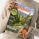 Eine hell gekleidete Person hält eine Ausgabe des Magazins „outdoor – Wandern. Reisen. Abenteuer.“ vor neutralem Hintergrund. Auf dem Titelbild: eine Berglandschaft mit grünen Wäldern, Felsen und Wandernden auf einem Pfad. Sichtbare Texte u. a.: „TOP-EQUIPMENT FÜRS CAMPING“, „Bayerns stille Winkel“, „Abenteuer Jotunheimen“, „Schwarzwald für Entdecker“ und „Im Check: Softshells“.