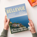 Bellevue-Sonderausgabe mit blauem Cover, gelber Titelzeile „BELLEVUE“ und dem Text „Handverlesene Domizile – von der Promi-Villa bis zur Karibik-Insel“ sowie „Wohnträume zwischen Frankfurt, Florida und Fidschi“. Zwei Hände einer nicht näher bestimmbaren Person halten das Magazin über einer weißen Fläche; das Cover zeigt Meer, Küste und Villen, oben rechts liegt ein orangefarbenes Notizbuch.