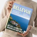Person in beigefarbenem Strickpullover hält eine schräg ausgerichtete Zeitschrift mit blau-gelbem Cover. Sichtbare Texte: „BELLEVUE“, „SONDERAUSGABE“, „HANDVERLESENE DOMIZILE – VON DER PROMI-VILLA BIS ZUR KARIBIK-INSEL“, „WOHNTRÄUME ZWISCHEN FRANKFURT, FLORIDA UND FIDSCHI“ und „FÜR UNSERE LESER AUSGESUCHT: DIE 300 SCHÖNSTEN IMMOBILIEN DER WELT“; darunter eine Küstenvilla am Meer.