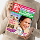 Weibliche Person in beigem Strickpullover hält eine schräg liegende Zeitschrift mit rotem Titel „Bild der Frau“. Das Cover zeigt eine lächelnde Frau sowie die Texte „Das dürfen Nachbarn jetzt“, „Drohnen, Kameras, Balkon-Kraftwerke“, „Wie schalte ich auf schlanke?“, „Neue Frisuren“ und „Pfefferminze hilft“.